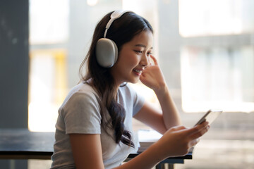 Fototapeta premium Asian teenage student woman wearing headphone to listening music and surfing social media on smartphone to relaxation with happiness after studying education knowledge for preparing test exam in cafe