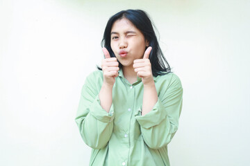 good job, very good. pleased asian woman wearing green over size shirt showing thumbs up with two hands and flirting to camera with blink eye closed standing over isolated white background