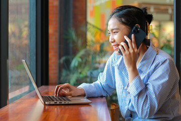 excited asian woman sitting in cafe bar working on laptop and talking on mobile phone