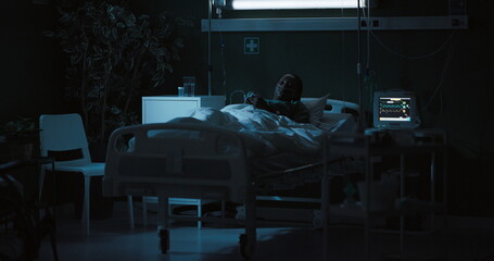 African woman in a hospital room lying on a bed chatting online with friends smiling.