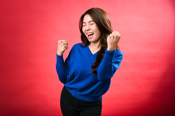 An Asian woman in a blue sweater and black pants stands against a red background with her eyes closed, fists clenched, and an excited expression, as if celebrating a victory.