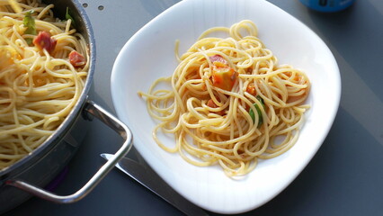 Portion of spaghetti served on a white plate next to a pan of freshly cooked pasta. The setting is casual, perfect for a relaxed outdoor meal with family and friends