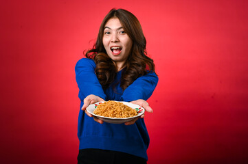 An Asian woman wearing a blue sweater is enthusiastically holding out a plate of noodles toward the...