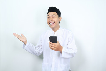 pleased asian muslim man smiling while presenting hand to beside with holding mobile phone isolated