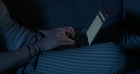 A man uses a laptop while sitting on the couch at night. Close-up of his hands, an unrecognizable person