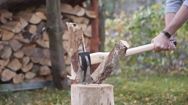 Wood is split with a sharp axe on a stump, filmed in slow motion. Wood chips fly through the air. A sunny garden scene with firewood in the background.