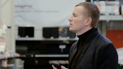 Man contemplating building materials for apartment renovation while holding smartphone in hardware store. A man in a hardware store, choosing flooring, shelves with ceramic tiles.