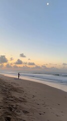 A lone surfer stands on a sandy beach at dusk, facing the ocean, with a crescent moon in the sky.