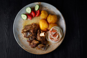 fried lamb ribs, meat with baked potatoes in oil with fresh tomatoes, cucumbers and onions on a black wooden background