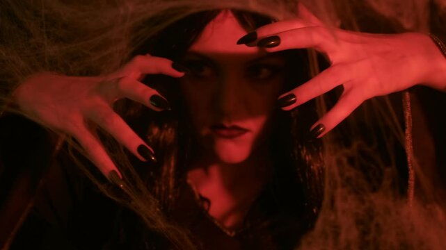 A dark-haired witch, with dramatic makeup, moves her hands as if casting a spell in warm, candlelit surroundings, with spider webs in the foreground.