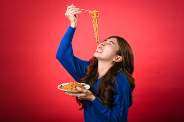 An Asian woman in a blue sweater excitedly lifts noodles with fork, ready to eat from the plate she...