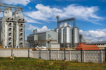 Obraz premium large grain elevator with multiple cylindrical metal silos, structural elements, and a complex system of pipes. It stands against a blue sky, with a field in the foreground after harvest