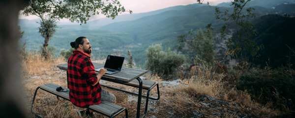 A young man is working on a laptop at a camping site in the mountains.