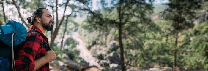 A male tourist with a large backpack and equipment on a mountain trail among the trees.