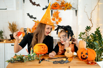Halloween, a happy family a mother with a baby girl in festive costumes and makeup is preparing for Halloween at home in the kitchen by coloring and carving pumpkins and scaring everyone