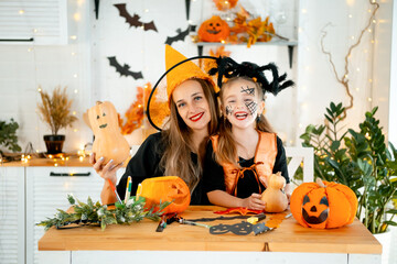 Halloween, a happy family a mother with a baby girl in festive costumes and makeup is preparing for Halloween at home in the kitchen by coloring and carving pumpkins and scaring everyone