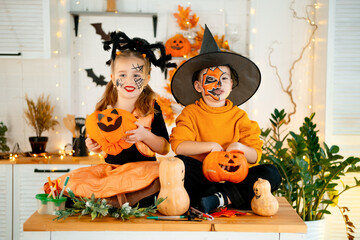 Halloween, two baby boy and girl in festive costumes and makeup are preparing for Halloween at home in the kitchen by coloring and carving pumpkins and scaring everyone
