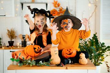 Halloween, two baby boy and girl in festive costumes and makeup are preparing for Halloween at home in the kitchen by coloring and carving pumpkins and scaring everyone