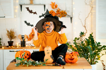 Halloween, a child boy in a festive costume and makeup is preparing for Halloween at home in the kitchen by coloring and carving pumpkins and scaring everyone