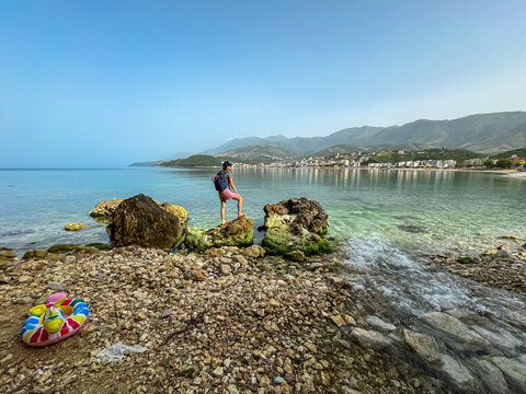 Woman on rock with panoramic view of coastline of tourist town Himare, Vlore, Albania. Sand beach Potami with scenic vistas of majestic Ceraunian mountain range. Summer vacation at Mediterranean sea
