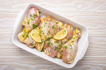 Raw uncooked chicken legs marinated with seasonings, herbs, lemon in white ceramic casserole top view on light wooden rustic background. Preparing healthy meal with marinated chicken drumsticks.
