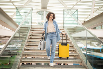 A woman with curly hair walks down the stairs carrying a yellow suitcase and a bag. She smiles...