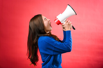 An Asian woman wearing a blue sweater is holding a red and white megaphone while speaking into it. She appears to be shouting or making an announcement. The background is solid red