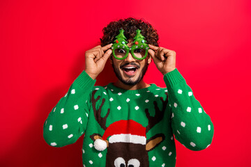 Portrait of nice young man touch tree shape glasses wear new year xmas sweater isolated on red color background
