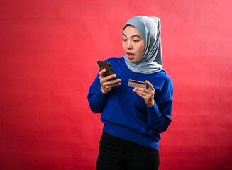 An Asian woman wearing a gray hijab and blue sweater looks excited while holding a smartphone in...