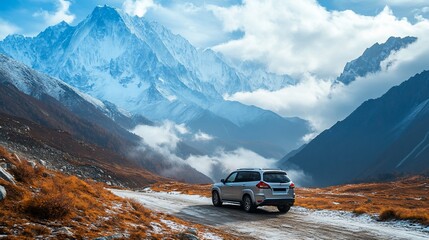 A silver SUV drives on a dirt road winding through a mountain valley with snow-capped peaks in the background.