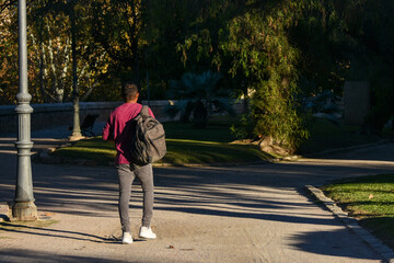 A young Hispanic man wearing a sweater and carrying a backpack walks in a park during the afternoon