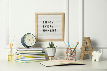 Letter board with phrase Enjoy Every Moment, alarm clock, decor and stationery on white desk