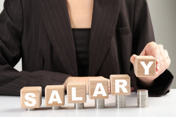 Woman putting wooden cubes with word Salary and stacks of coins on white table, closeup