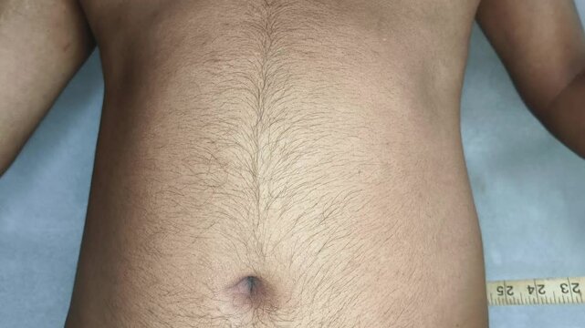 Indian or Asian young adult man holding a sewing meter, measuring his waist, abdomen, tummy or stomach and checking excessive belly fat. Overweight, obesity, weight loss and losing calorie concept.