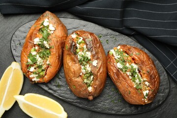 Tasty baked sweet potatoes with feta cheese, parsley and lemon slices on dark textured table, flat lay