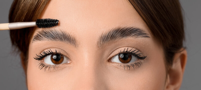 Young woman correcting her eyebrows on grey background, closeup