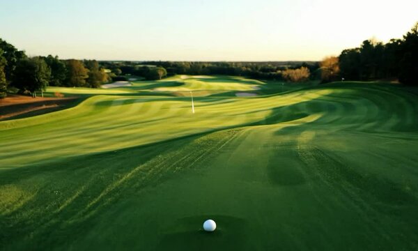 Tilt shot with early morning light: The camera starts with a close-up of a golf ball on the tee. It tilts up to reveal the golf course bathed in the soft, early morning light, highlighting the dew-cov