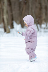 Side view toddler girl in light pink jumpsuit and a white fluffy hat walking in winter park. Children games in the snowy forest. Family winter vacation with a child. Vertical photo