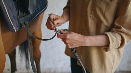 Equestrienne hands fastening stirrup on horse at paddock closeup. Woman animal