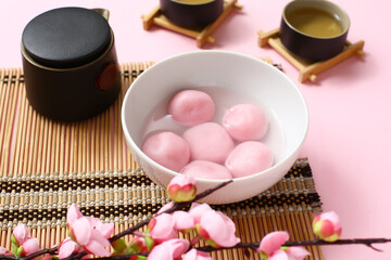 Bamboo mat with bowl of tangyuan and sakura on pink background, closeup. Dongzhi Festival