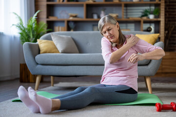 Senior woman with shoulder pain sits on yoga mat in living room. Elderly lady in pink shirt and...