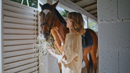 Positive woman feeding equine light barn closeup. Lady equestrienne giving hay © stockbusters
