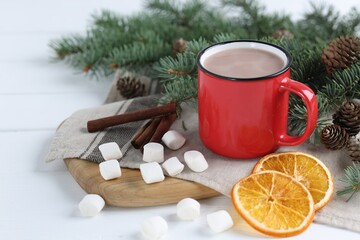 Yummy cocoa in red cup, marshmallows, cinnamon sticks, dried orange slices and fir tree branches on white wooden table. Space for text