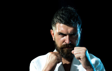 Portrait of strong karate man in kimono at gym. Bearded male practicing aikido. Handsome muscular fighter ready for karate training. Serious karate man in fighting position preparing for competition.