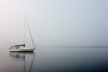 Fototapeta premium Sailboat rests moored in the calm harbor waters in morning fog at Egg Harbor, Door County, Wisconsin in late spring