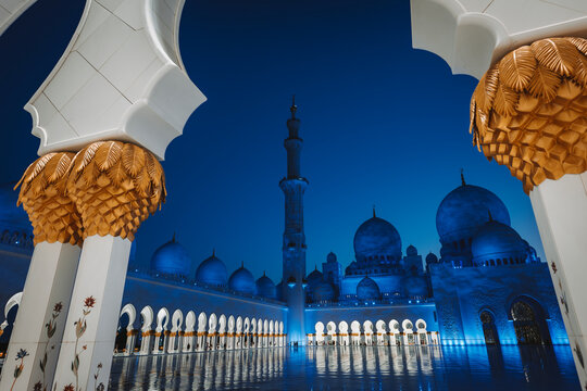 Night view of the Sheikh Zayed Grand Mosque in Abu Dhabi, illuminated in blue, with ornate columns, domes, and reflections across the polished marble courtyard.