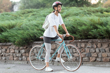 Naklejka premium Happy young woman in helmet and casual clothes riding bicycle outdoors, enjoying healthy lifestyle and urban commute, green bush and a stone wall in the background