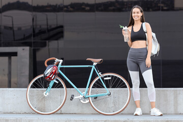 Sporty young woman with bicycle and bottle of water on city street