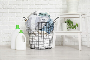 Basket with dirty clothes, bottles of laundry detergent and houseplant near white brick wall