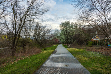 Wet paved walkway through spring park with bare trees and budding blossoms under cloudy sky. New Jersey. USA.